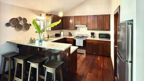 Une cuisine moderne avec des meubles de couleur sombre en bois, un réfrigérateur en acier inoxydable, un four à micro-ondes et une cuisinière à gaz. Il y a trois tabourets au comptoir central.