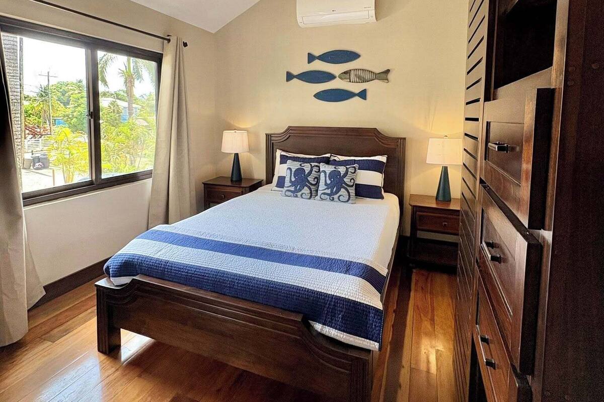 A bedroom with a bed, two bedside tables with lamps, a ceiling fan, a wall-mounted air conditioner, and a window with curtains.