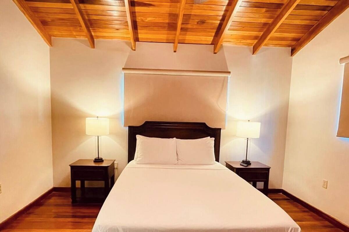 A bedroom with a wooden ceiling, a large bed with white bedding, two bedside tables with lamps, and a window on the right wall.