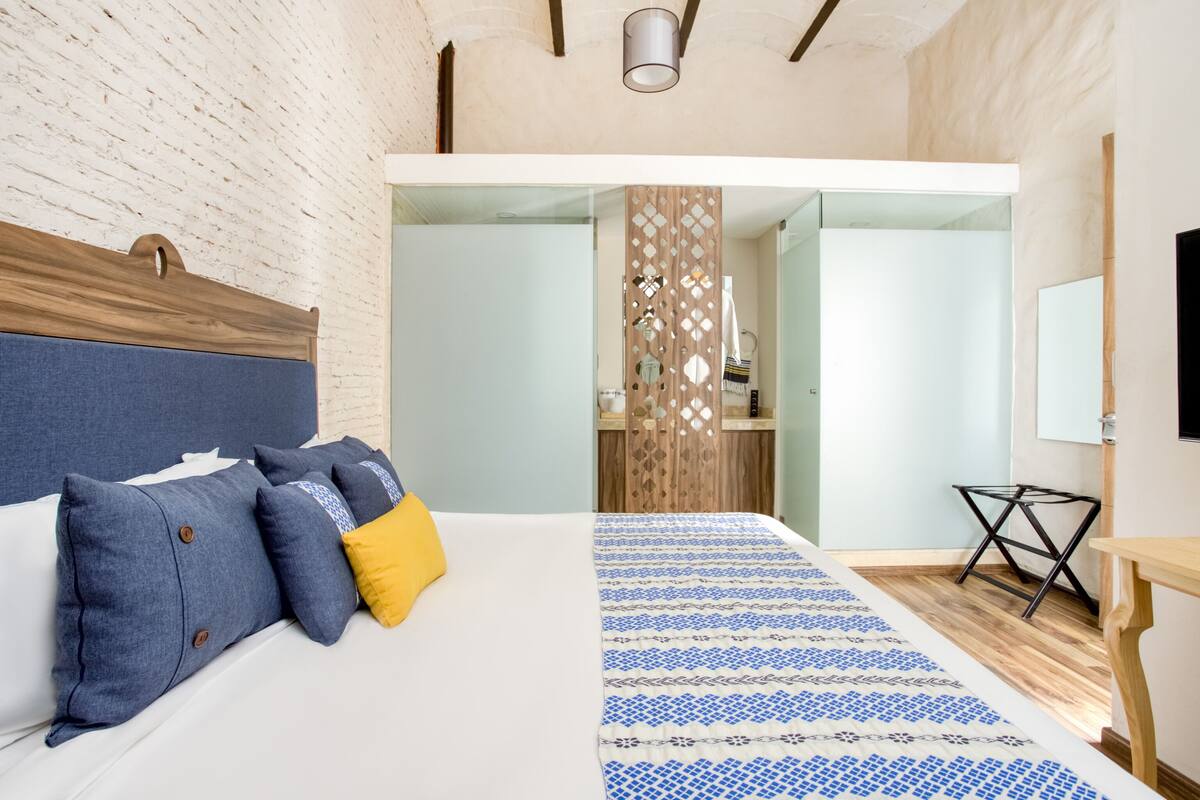 A modern hotel room with a large bed, a wooden headboard, a patterned bedspread, a small table, and a mirror on the wall.