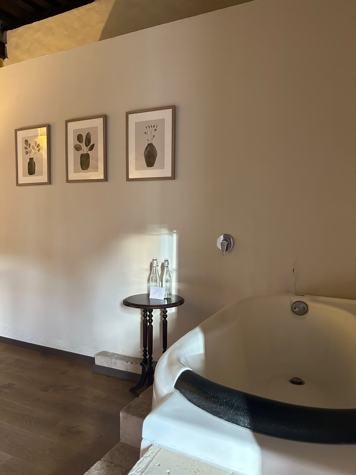 A bathroom with a large bathtub, a small table with bottles, and three framed pictures on the wall.