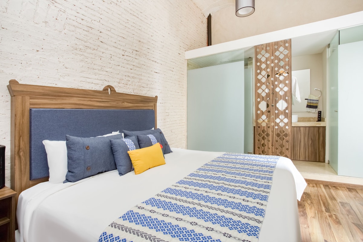 A modern bedroom with a wooden headboard, a patterned bedspread, and a view of a bathroom with a wooden partition.