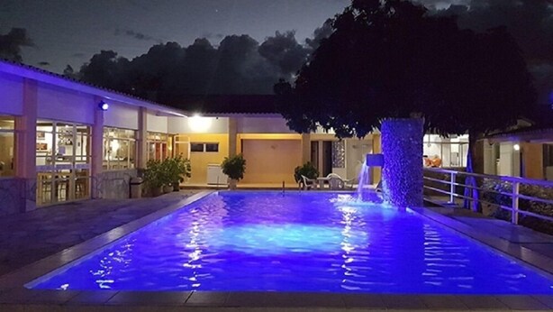 Una piscina iluminada en azul, un árbol y un edificio con grandes ventanales.