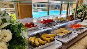 Un bufé con diversas frutas y verduras, incluyendo plátanos, manzanas y sandía, en una sala con vista a la piscina.