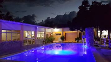 Piscina iluminada em azul, área de varanda coberta e um edifício com amplas janelas e interior visível.