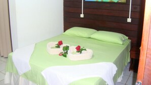 A neatly made bed with a green bedspread and white linens, a small white table with a red flower, and a painting of a coastal scene on the wall. - Pousada Raio de Sol (Fernando de Noronha)