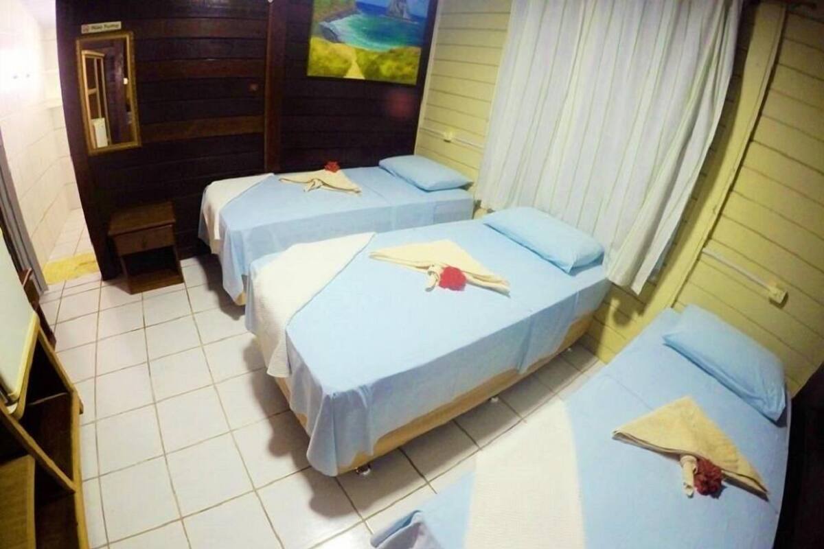 Ein Hotelzimmer mit drei Betten, jeweils mit blauer Bettdecke und weißen Kissen. Es gibt ein hölzernes Kopfteil, ein Bild an der Wand und ein Fenster mit weißen Vorhängen.