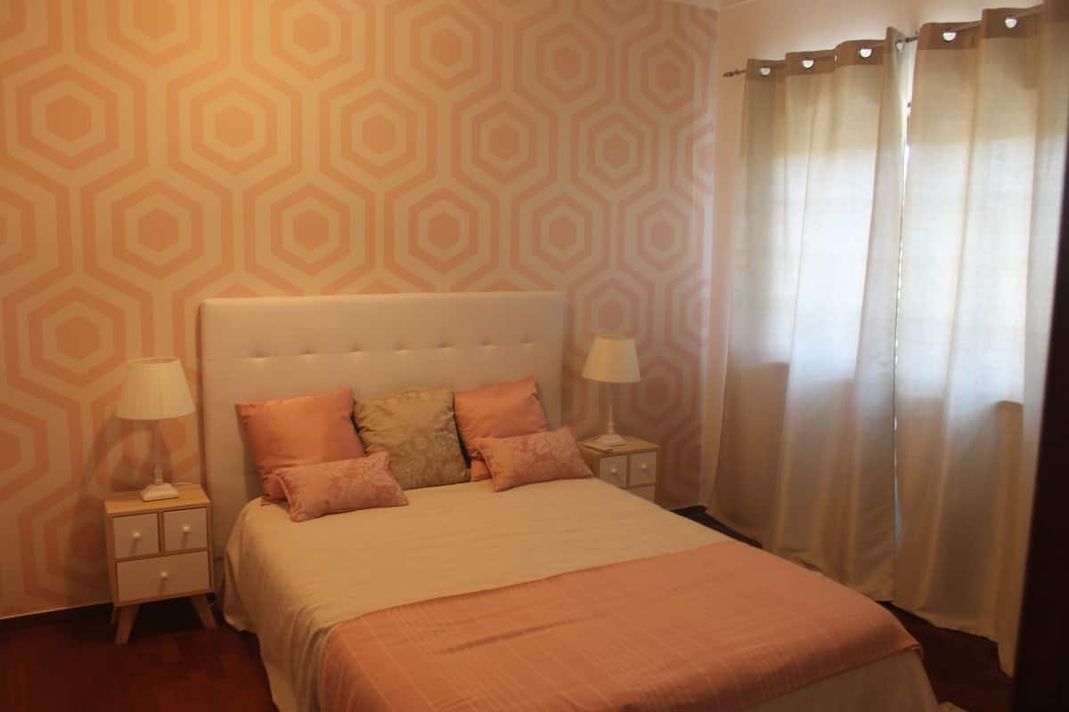 A neatly arranged bedroom with a large bed, two bedside tables with lamps, and a window with sheer curtains.