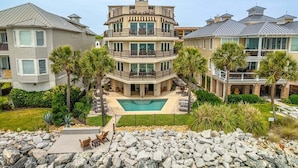 A multi-story residential building with a swimming pool, surrounded by palm trees and a rocky landscape.