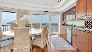 A dining area with wicker chairs and a round table, wooden cabinets, and a view of the ocean through large sliding doors.