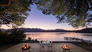 A wooden deck with a table and chairs, a fire pit, and a view of a calm lake with mountains in the background. - Calfuco Wine Hotel & Spa (Villa La Angostura)