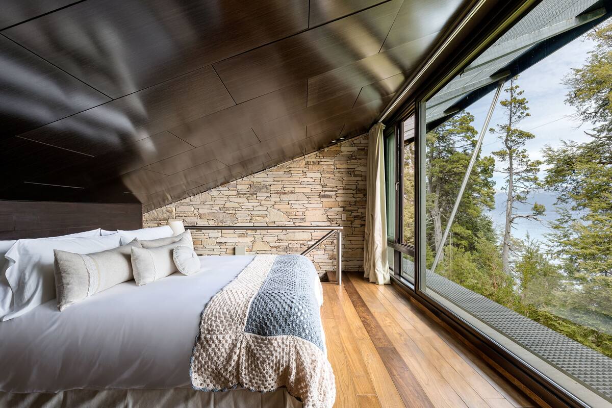 Ein Schlafzimmer mit Steinsandsteinwand, einem großen Bett mit Kissen, einer Decke und Blick auf Bäume durch ein Fenster.