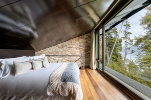 A bedroom with a stone wall, a large bed with pillows, a blanket, and a view of trees through a window.