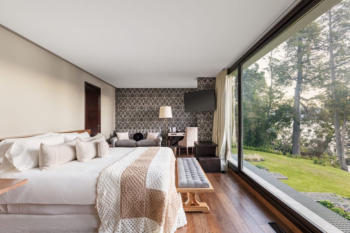 Ein Schlafzimmer mit einem großen Bett, einem Schreibtisch, einem Stuhl und Blick auf einen Wald durch ein großes Fenster.