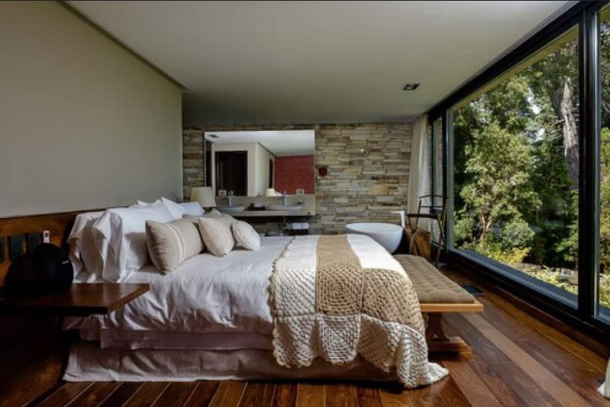 Ein modernes Schlafzimmer mit einem großen Bett, einer hölzernen Bank, einer Steinwand und Blick auf Bäume durch große Fenster.