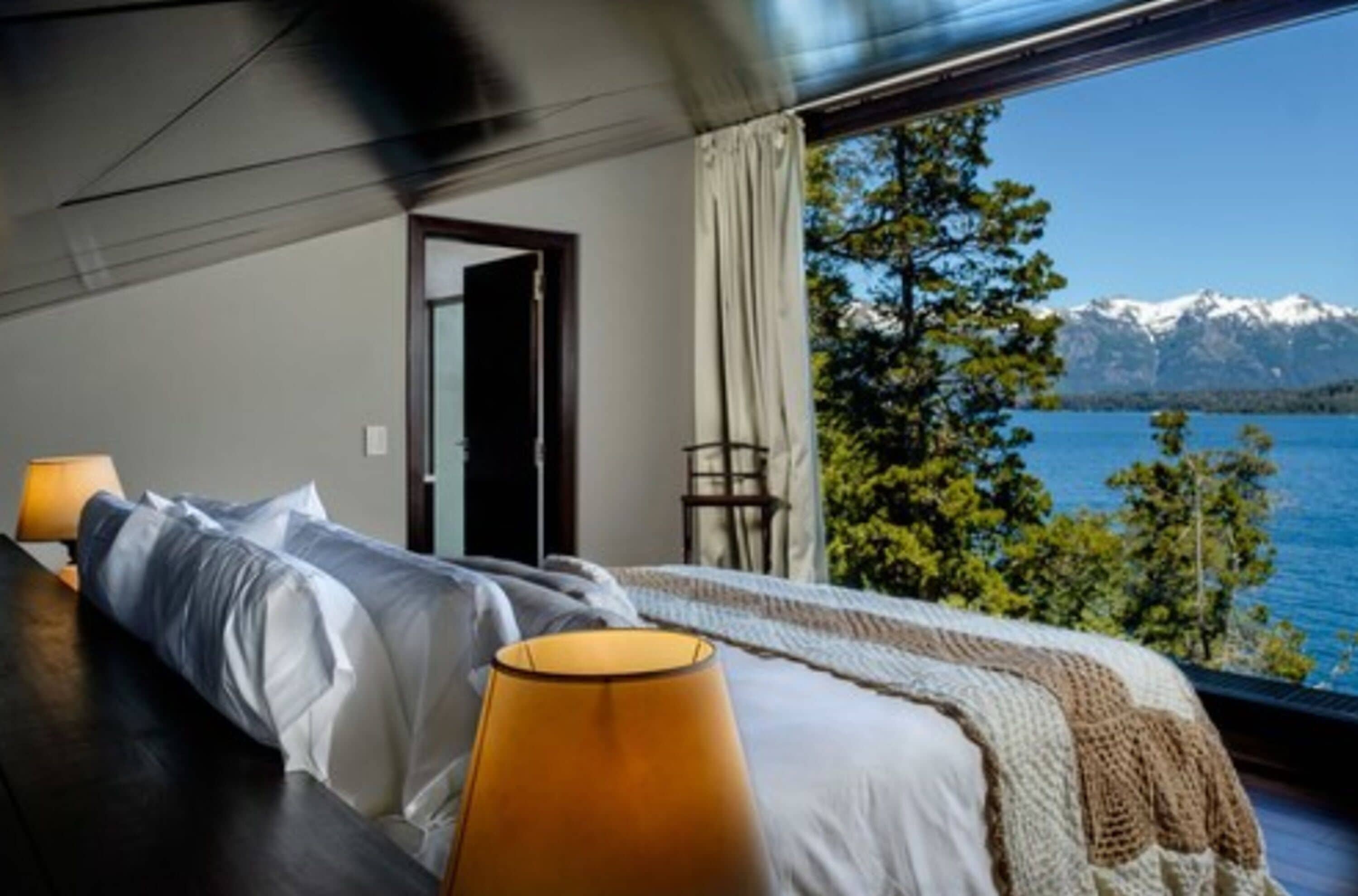 A bedroom with a bed, a lamp, and a view of a lake and mountains.
