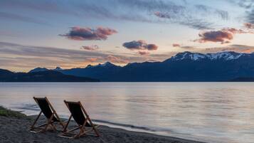 Una playa con dos sillas de madera mirando un lago tranquilo y montañas nevadas al fondo.