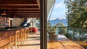 A wooden deck with a bar area, wicker chairs, and a table with a view of mountains and trees. - Calfuco Wine Hotel & Spa (Villa La Angostura)