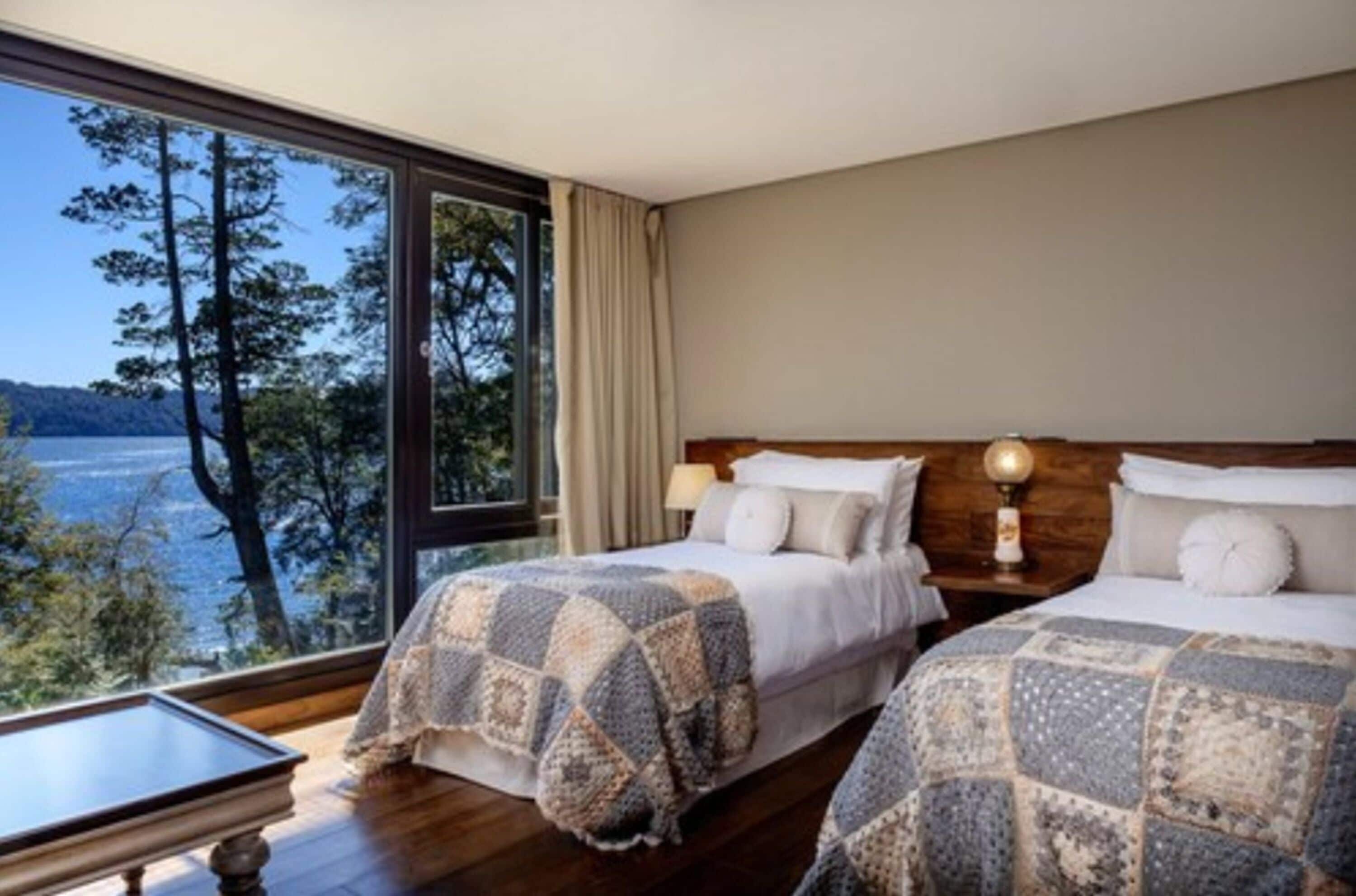 Un dormitorio con dos camas, una mesita de noche con lámpara y vistas a un lago a través de amplias ventanas.