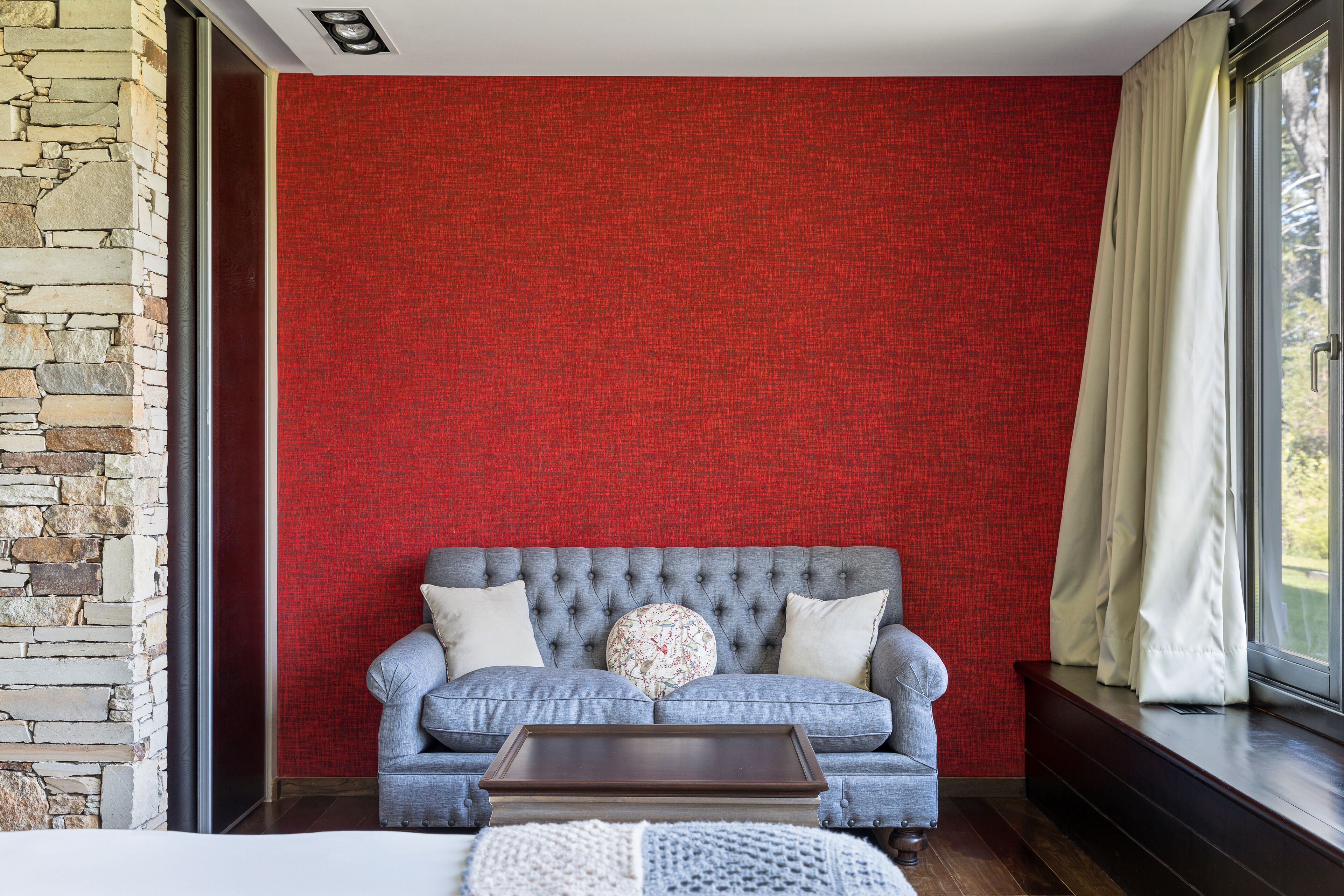 A room with a stone wall, a red textured wall, a grey sofa with cushions, a wooden coffee table, and a window with curtains.