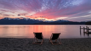 Two beach chairs facing a calm lake with a sunset and mountains in the background. - Calfuco Wine Hotel & Spa (Villa La Angostura)
