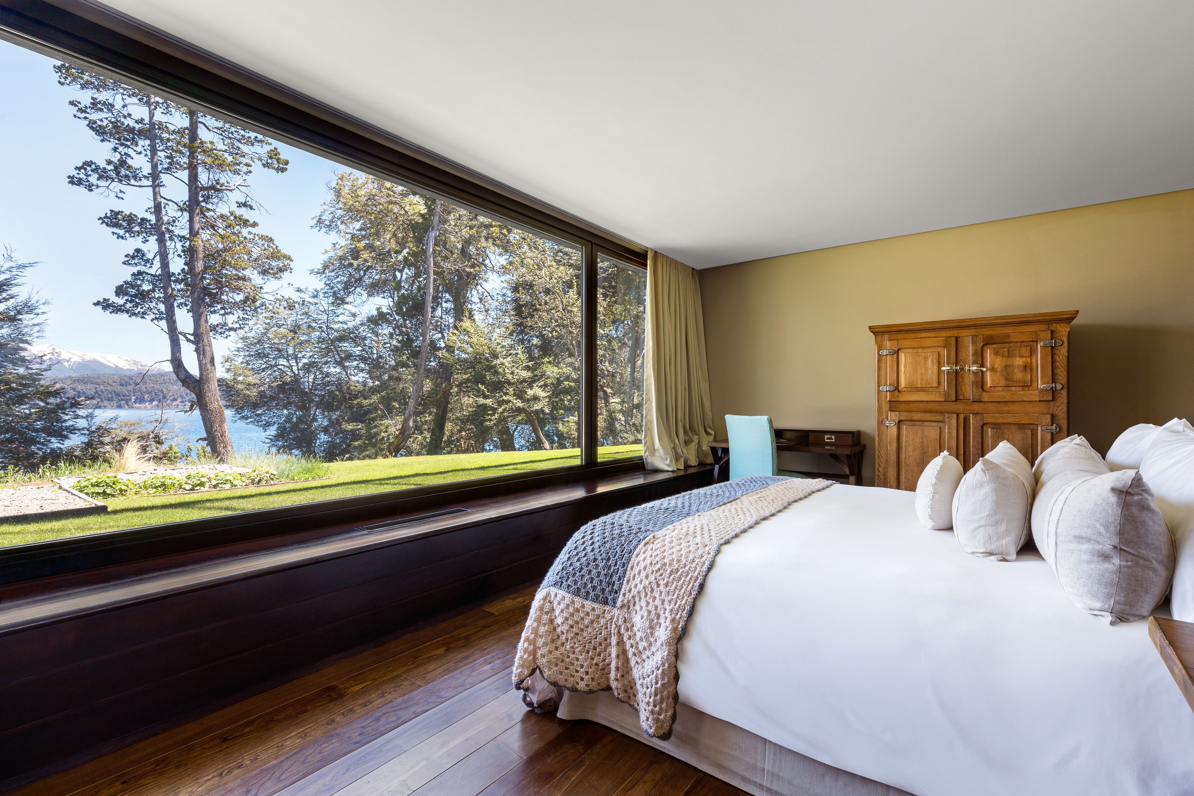 A bedroom with a large window, a bed with white bedding, a wooden wardrobe, and a view of trees and mountains.