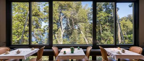 A dining area with a table set for four, large windows offering a forest view, and wooden flooring.