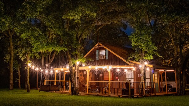 Una cabaña de madera con un porche, rodeada de árboles e iluminada por luces colgantes.