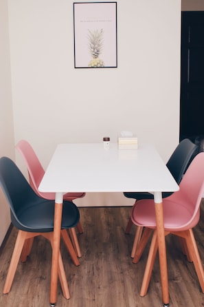A white table with a framed botanical print above it, surrounded by four chairs with wooden legs and colorful upholstery.