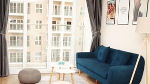 Ein modernes Wohnzimmer mit einem blauen Sofa, einem runden weißen Hocker, einem kleinen weißen Tisch mit zwei Flaschen und einem großen Fenster mit Vorhängen.