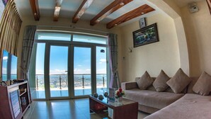 A living room with a sofa, a coffee table, and a television. There is a large window with a view of the outdoors. - Ho May Resort (Ho Chi Minh City)