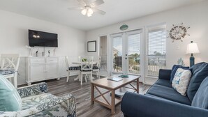 A living room with a TV, ceiling fan, white furniture, and a view through the glass doors.