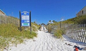 A sandy pathway leading to a beach area with a 'Safety First' sign, a wooden fence, and a small structure in the distance.