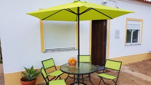 En udendørs patio med et rundt glasbord, fire grønne stole, en gul parasol og en potteplante.