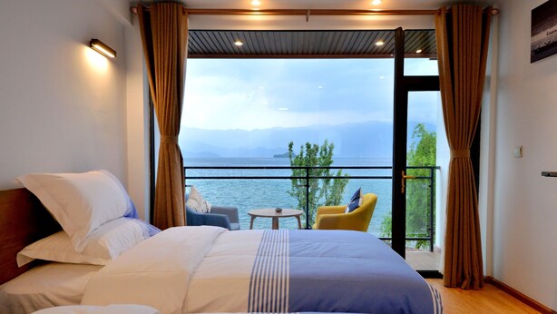 Ein Schlafzimmer mit einem großen Bett, einem kleinen Tisch und einem Stuhl, inklusive Balkon und Blick auf Wasser und Berge.