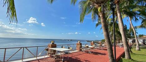 A seaside patio with tables and chairs, palm trees, and a view of the ocean.