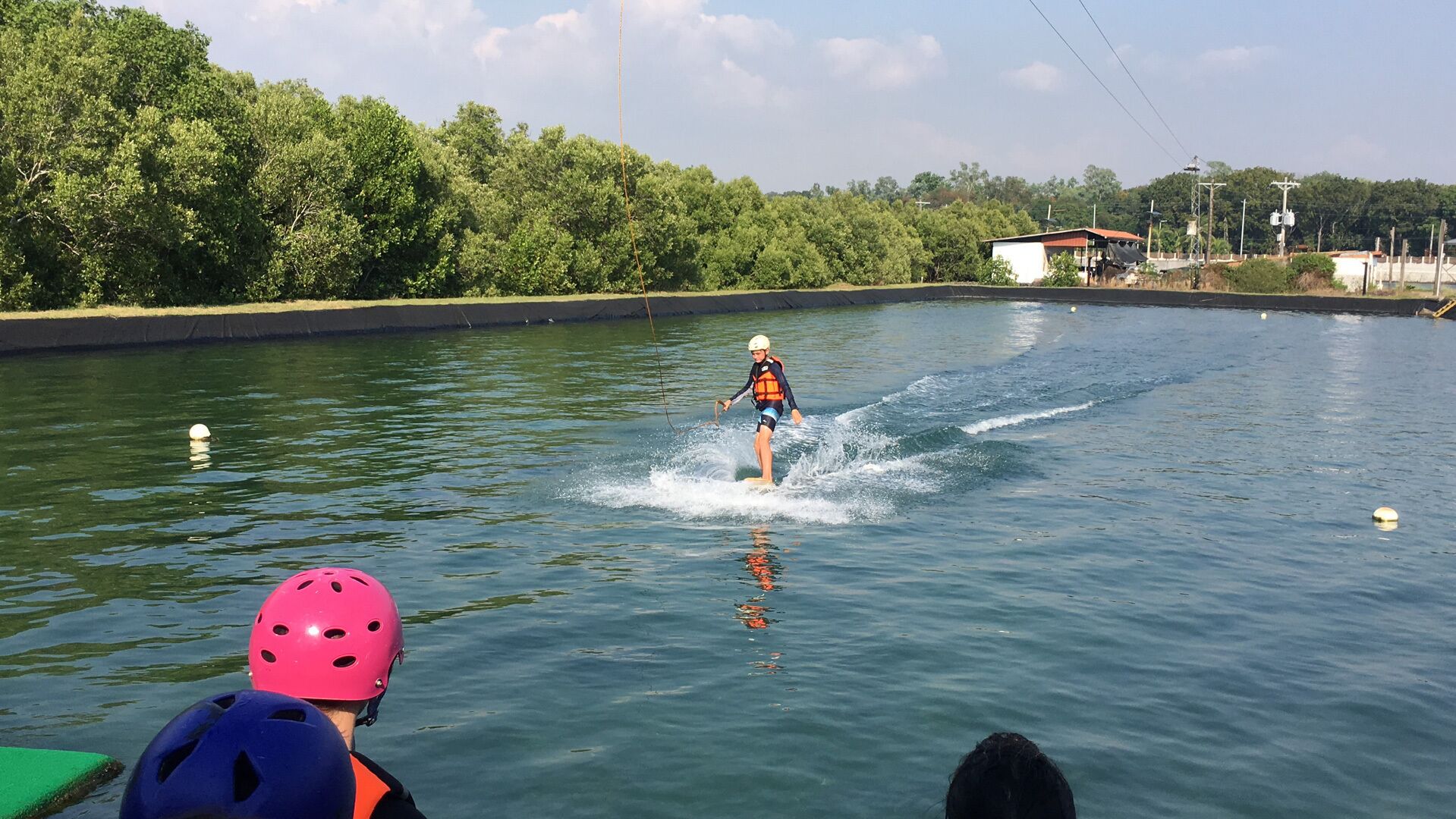 En person laver wakeboard på stille vand, omgivet af grøn vegetation og nogle bygninger i baggrunden.