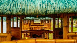 Una estructura de madera con techo de paja, posiblemente una cabaña o un pabellón, con vistas a un cuerpo de agua y follaje al fondo.
