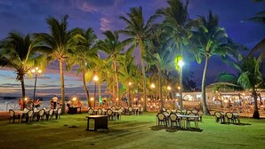 Un área de comedor al aire libre con mesas y sillas, rodeada de palmeras, bajo luces colgantes.