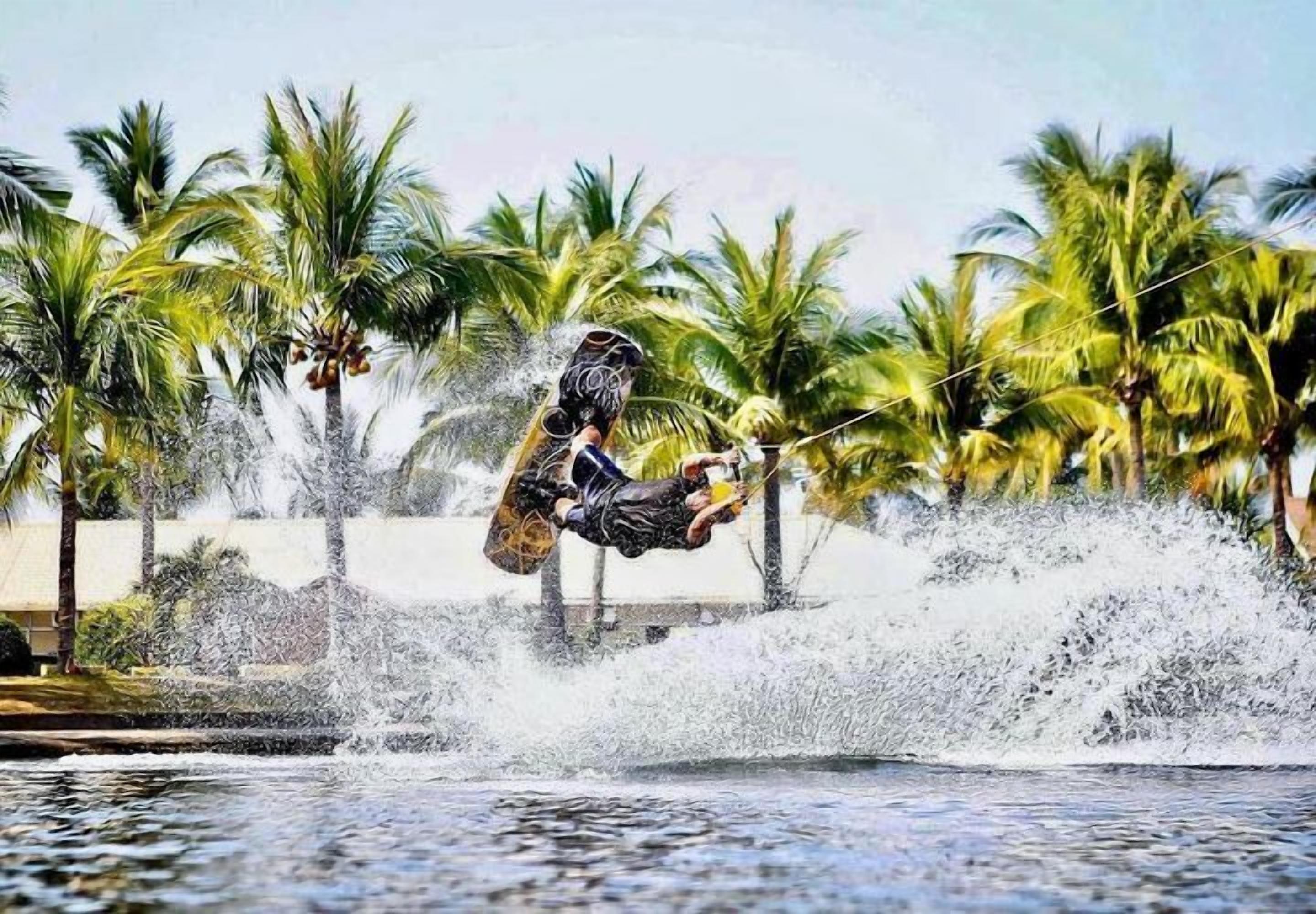 A wakeboarding stunt with a person mid-air over a body of water, surrounded by palm trees.