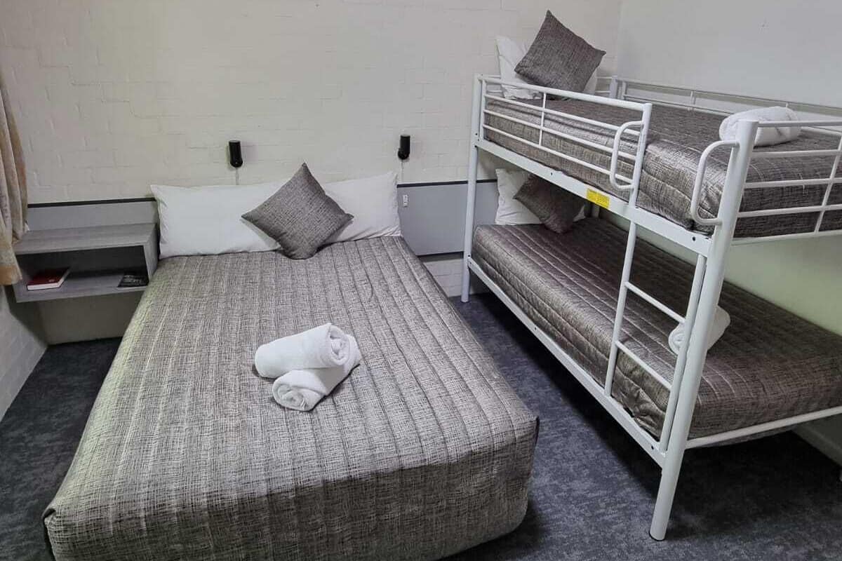 A single bed with a grey mattress and pillows, a white headboard, a bedside table, and a white bunk bed to the right.