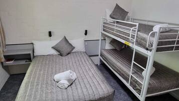Una cama individual con colchón y almohadas grises, cabecero blanco, mesita de noche y una cama nido blanca a la derecha.