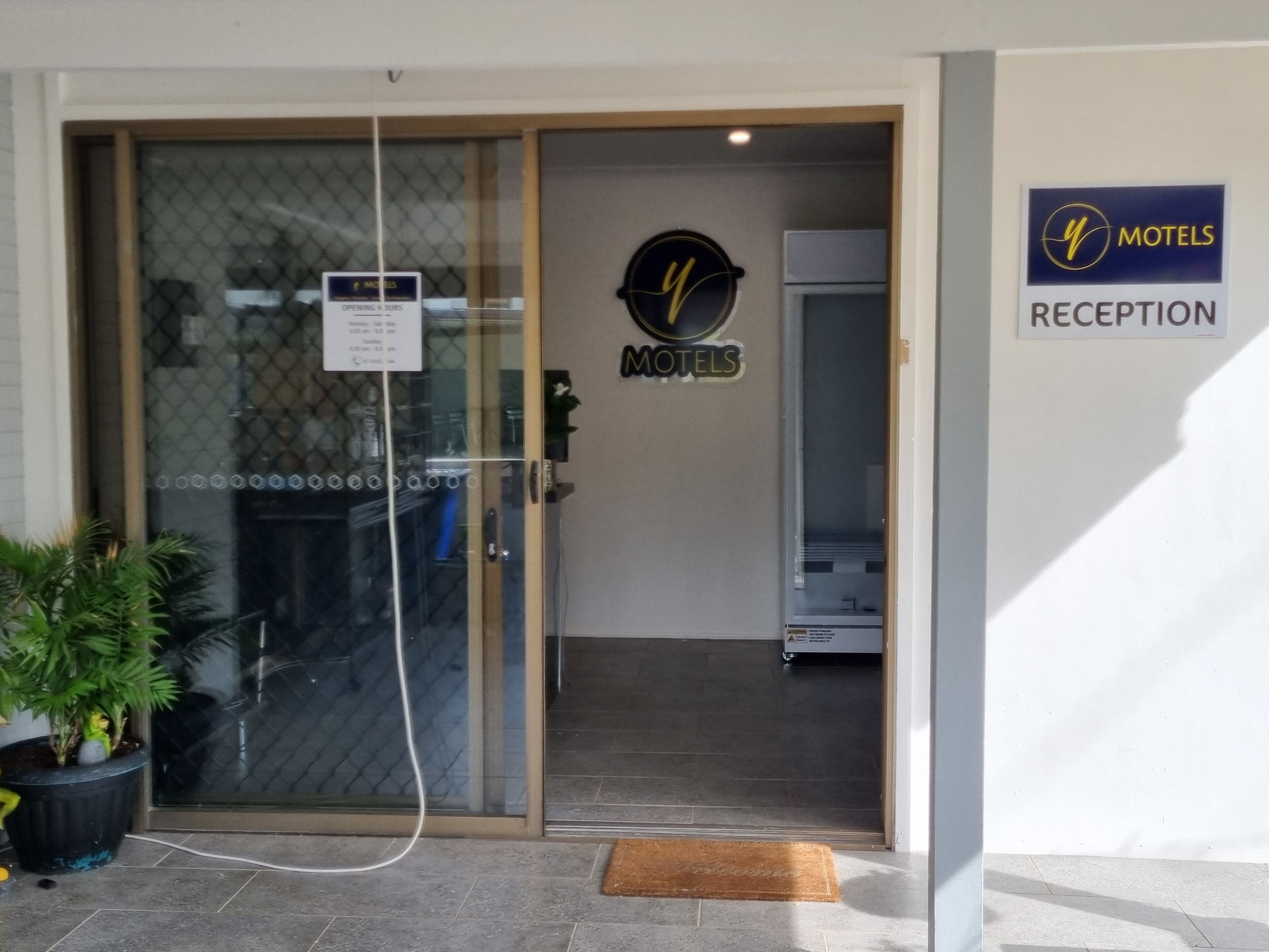 Un espace d’accueil dans un motel, avec une porte vitrée, un comptoir de réception et un panneau indiquant que la réception se trouve à cet endroit.