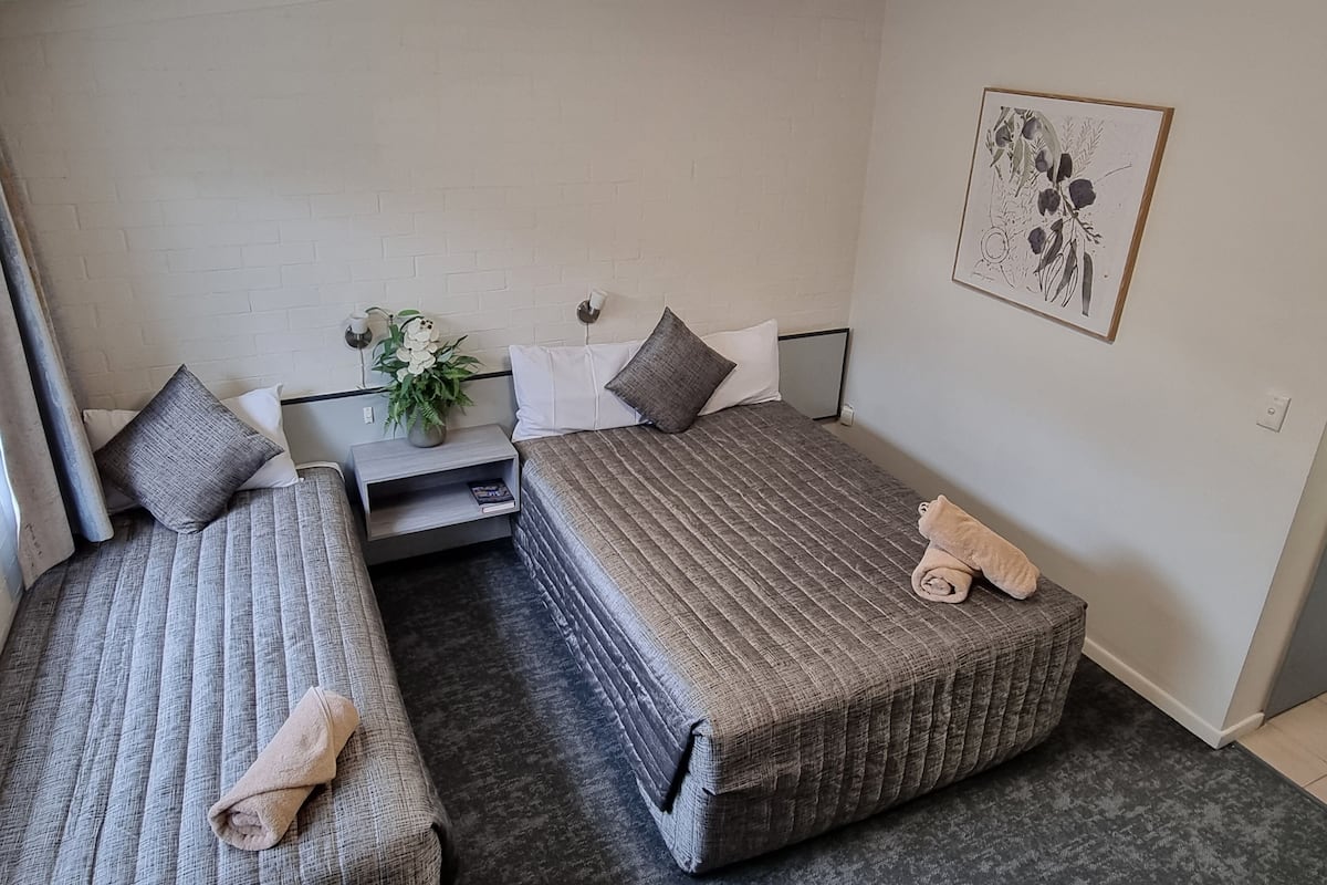 A small, clean room with two single beds, a bedside table with a lamp, a small plant, and a framed picture on the wall.