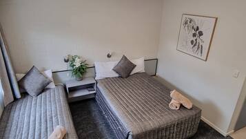 A small, clean room with two single beds, a bedside table with a lamp, a small plant, and a framed picture on the wall.