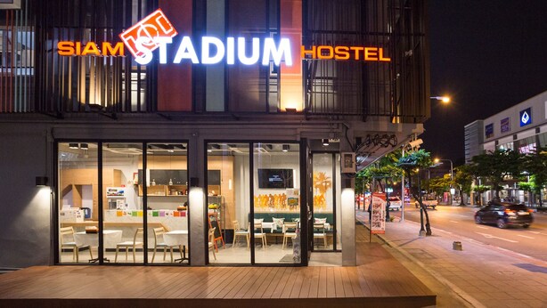 Ein Hotel namens "SIAM STADIUM HOSTEL" mit modernem Design, einer Glasfassade, einem Sitzbereich im Freien und einem gut beleuchteten Eingang.
