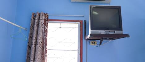 A room with a blue wall, an air conditioner, a television, and a window with patterned curtains.
