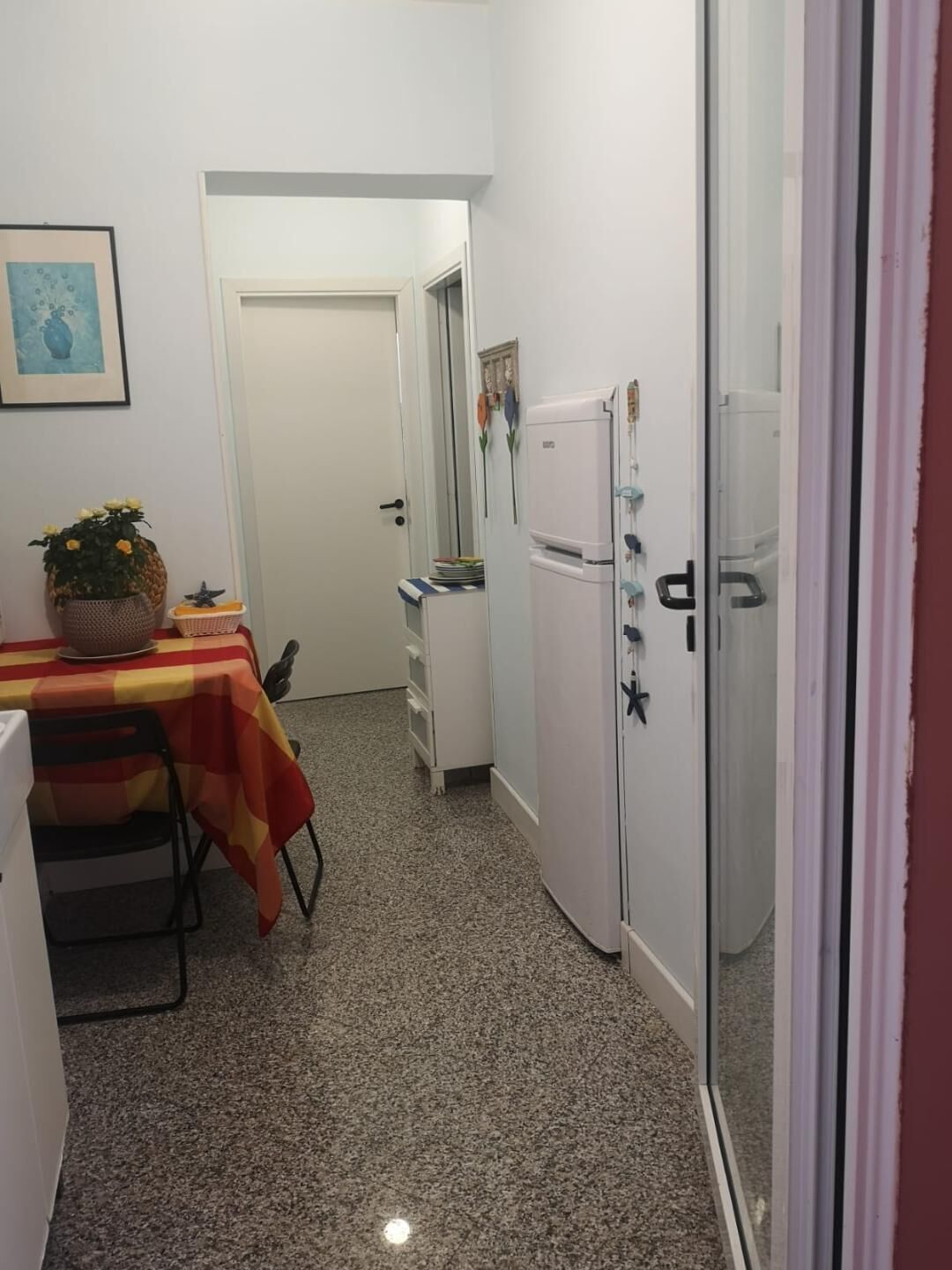 A compact kitchen with a refrigerator, a small dining table with a red and yellow tablecloth, and a potted plant.