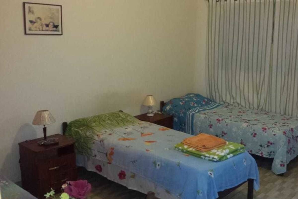 A bedroom with two beds, each with a nightstand and lamp. There is a window with vertical blinds, and a framed picture on the wall.