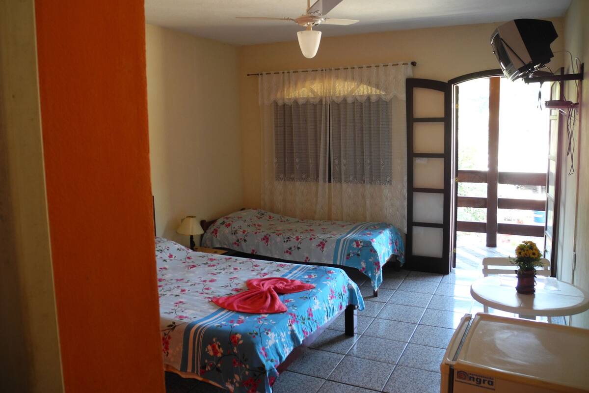A room with two beds, a ceiling fan, a window with curtains, and a small table with a flower vase.